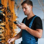 Young man in uniform with measuring device works with internet equipment and wires in server room.